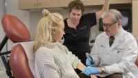 A woman sits in an examination chair and has her skin inspected by a doctor. A nurse stands in the background holding a light over them